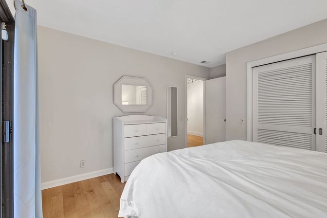Bright minimalist apartment bedroom with white bedding, white dresser under an octagonal mirror, light hardwood floors, sliding louvered closet doors and a balcony curtain