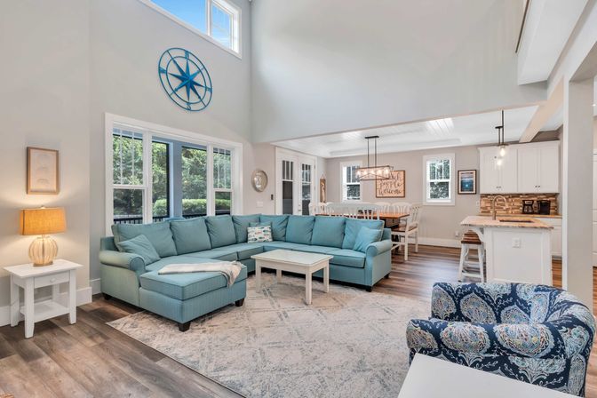 Bright open-concept coastal living room and kitchen with teal sectional sofa, patterned accent chair, white coffee table, large windows, vaulted ceiling, dining area and island on wood-look floors.
