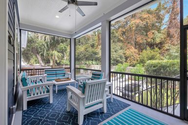 Bright screened porch with white wooden patio furniture and teal-striped cushions around a coffee table, blue patterned rugs, ceiling fan overhead, and views of a lush backyard with palm trees and a grill.