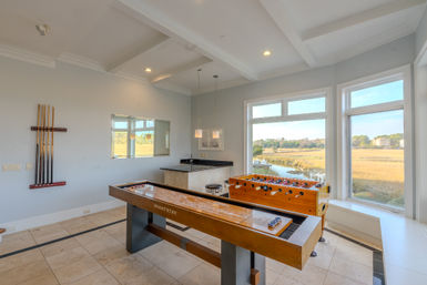 Sunlit waterfront game room with wooden shuffleboard and foosball tables, wall-mounted pool cues, small wet bar, and large windows overlooking a coastal marsh and river view.