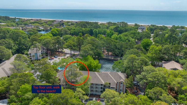 Aerial view of a sunny coastal condominium complex tucked into pine trees with the ocean on the horizon; a red circle highlights a top-floor unit, with on-site tennis courts, a pool, and a short walk to the beach.