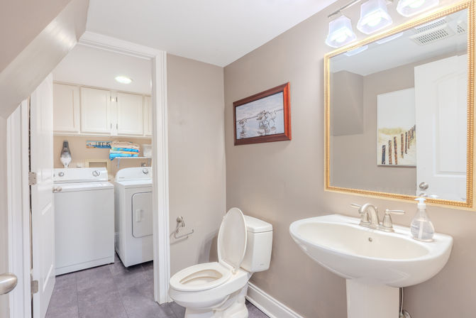 Cozy home half-bath with pedestal sink, toilet, gold-framed mirror and beach artwork; open doorway reveals laundry nook with white washer and dryer and overhead cabinets.