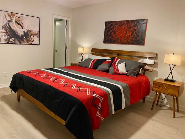 Cozy modern master bedroom with king-size bed in red, black and gray striped bedding, wooden slatted headboard, mid-century nightstands with lamps, abstract wall art and light hardwood floors.