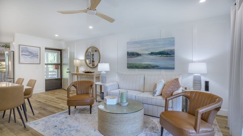 Bright modern coastal living room with white striped sofa, two caramel leather chairs, round woven glass-top coffee table, seascape painting, hardwood floors and a wooden ceiling fan.