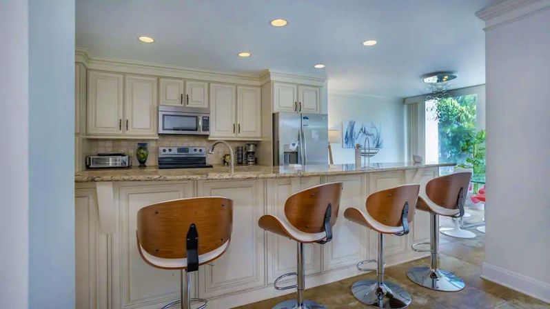 Sunny modern open-plan kitchen with cream cabinetry and granite breakfast bar, four curved wood-and-chrome bar stools, stainless steel appliances and a leafy window view.