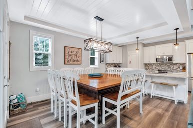 Bright beach-house dining area with a large wooden farmhouse table surrounded by white chairs, geometric pendant lighting and an open-plan kitchen with white cabinets, tile backsplash and breakfast bar.