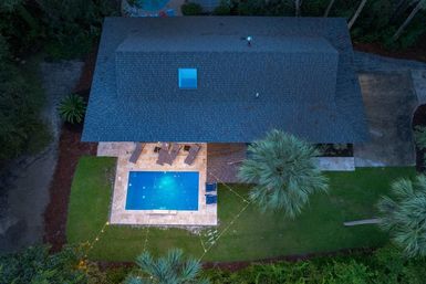 Aerial shot of a cozy suburban backyard at dusk: glowing rectangular pool on a tiled patio with lounge chairs, string lights, palm trees and green lawn.