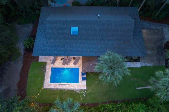 Aerial shot of a cozy suburban backyard at dusk: glowing rectangular pool on a tiled patio with lounge chairs, string lights, palm trees and green lawn.