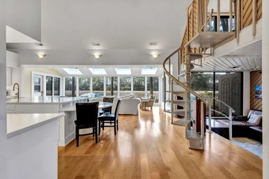 Sunlit open-plan waterfront living space with white kitchen island and bar seating, hardwood floors, metal spiral staircase, dining chairs, and a sunroom with skylights and floor-to-ceiling windows overlooking marsh and trees.