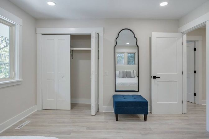 Bright modern bedroom interior with open white closet doors, arched black-framed wall mirror reflecting a bed, blue tufted ottoman on light wood floors, recessed ceiling lights and neutral walls.