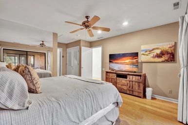 Coastal-inspired master bedroom with king bed and patterned pillows, hardwood floors, ceiling fan, wooden TV console showing a beach sunset and sliding patio doors
