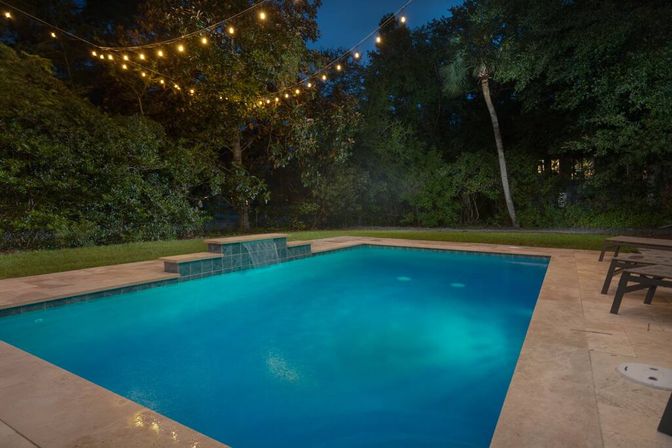 Nighttime backyard pool with glowing blue underwater lights, spillover waterfall feature, stone patio, lounge chairs and string lights over a wooded yard