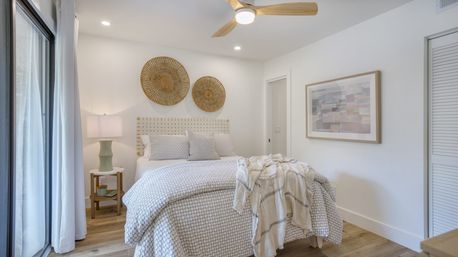 Light-filled modern coastal bedroom with patterned duvet, woven rattan wall art above the headboard, wood-blade ceiling fan, sliding glass doors and a bedside lamp