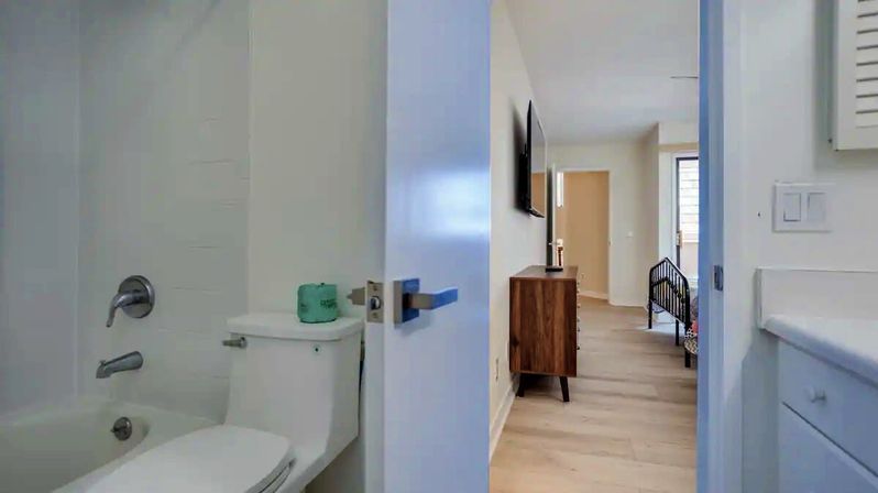 Bright modern white bathroom with tub, chrome fixtures and toilet; door opens onto light-wood hallway revealing a mid-century dresser, wall-mounted TV and a small bed in the adjacent room.