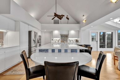 Bright modern white kitchen with vaulted ceiling, large island and round breakfast table with black chairs; stainless appliances, ceiling fan and sliding doors to a deck with water view.