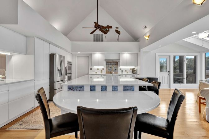 Bright modern white kitchen with vaulted ceiling, large island and round breakfast table with black chairs; stainless appliances, ceiling fan and sliding doors to a deck with water view.