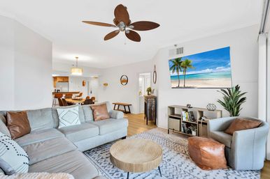 Bright coastal-style open-plan living room in a modern apartment, light-gray sectional with leather pillows, round wooden coffee table, swivel chair with leather pouf, ceiling fan, wall-mounted TV showing a tropical beach, dining area and kitchen visible in background.