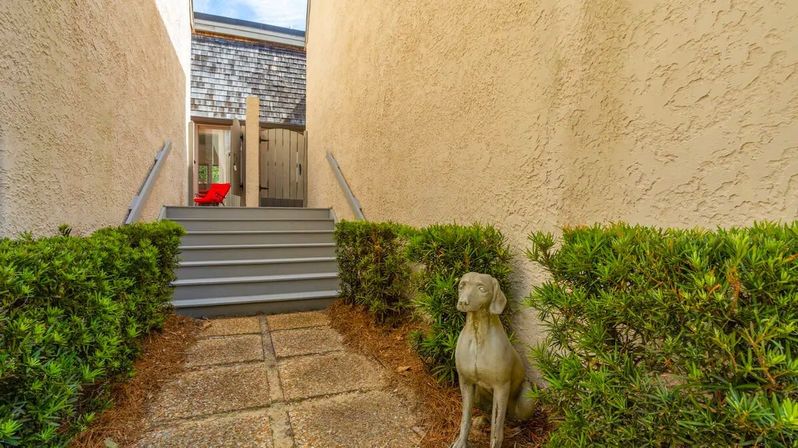 Narrow stucco-walled walkway with paver path and steps up to a small deck, trimmed hedges, a red patio chair, and a dog garden statue.