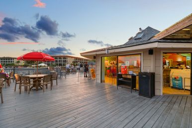 Sunset-lit waterfront boardwalk with outdoor dining on a wooden deck, red umbrella, and an open-air snack bar at a coastal resort