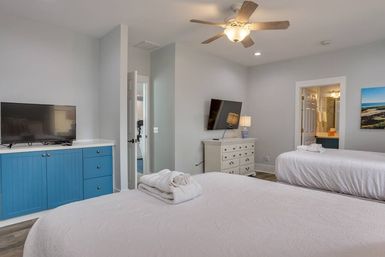 Bright, cozy modern guest bedroom with two beds in white linens and folded towels, blue built-in cabinet with TV, wall-mounted TV over a white dresser, ceiling fan, gray walls and open doorway to en-suite bathroom.