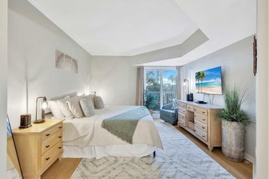 Sunny coastal-style master bedroom with king bed, neutral linens and green throw, light wood nightstand and dresser, wall-mounted TV and sliding glass doors opening to a balcony with palm-tree view