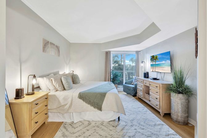Sunny coastal-style master bedroom with king bed, neutral linens and green throw, light wood nightstand and dresser, wall-mounted TV and sliding glass doors opening to a balcony with palm-tree view