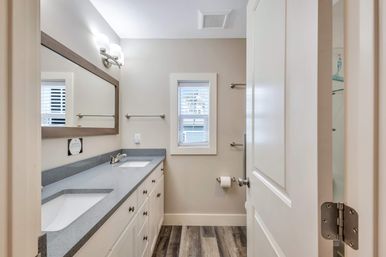 Bright modern residential bathroom with double sinks and gray quartz countertop, white shaker cabinets, large framed mirror, wall sconces, shuttered window, and wood-look plank flooring.