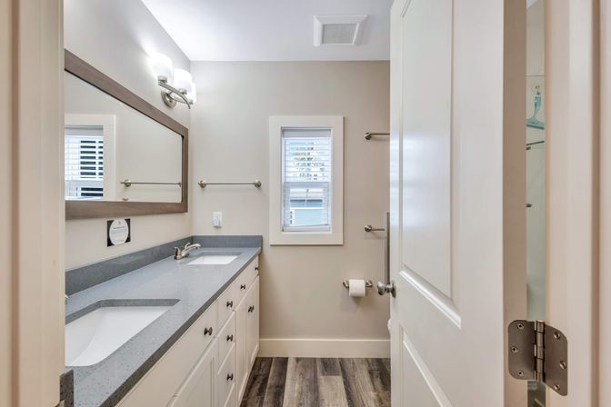 Bright modern residential bathroom with double sinks and gray quartz countertop, white shaker cabinets, large framed mirror, wall sconces, shuttered window, and wood-look plank flooring.