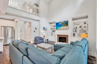 Bright coastal-style open-concept living room with vaulted ceiling and upstairs balcony, large blue sectional around a glass coffee table, brick fireplace with wall-mounted TV showing a beach, white built-in shelves, hardwood floors, and a stainless steel refrigerator visible to the kitchen.