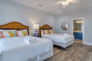 Bright guest bedroom in a vacation rental with two queen beds, wooden headboards, white linens and colorful pillows, folded towels on beds, blue-and-white lamp on a nightstand, round wall mirror, ensuite bathroom with blue vanity, ceiling fan and wood-look flooring.