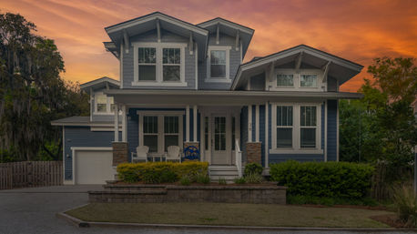Blue two-story Craftsman-style home with a covered front porch, white Adirondack chairs, attached garage and landscaped yard under a warm orange sunset sky.