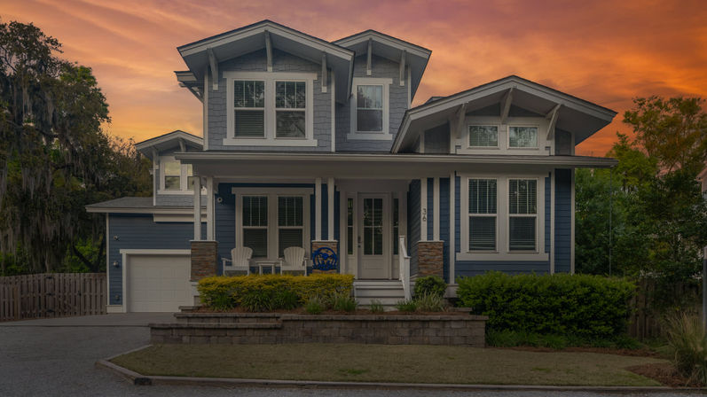Blue two-story Craftsman-style home with a covered front porch, white Adirondack chairs, attached garage and landscaped yard under a warm orange sunset sky.