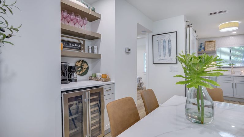 Bright modern coastal-style open-concept kitchen and dining area with built-in beverage bar, glass-front wine fridge, open shelves of glassware, tan leather chairs, and a vase of green foliage on a white marble table.