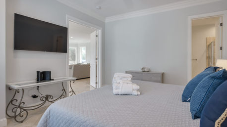 Sunlit modern bedroom with gray quilt and blue throw pillows, stacked white towels on the bed, wall-mounted TV above a decorative console table, and open doorways to a bright living area and bathroom.