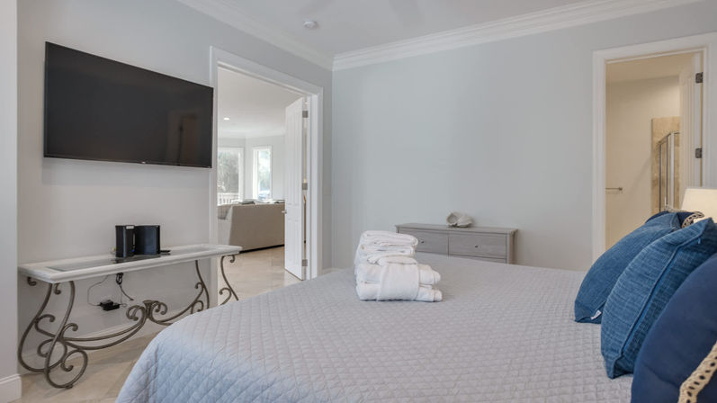 Sunlit modern bedroom with gray quilt and blue throw pillows, stacked white towels on the bed, wall-mounted TV above a decorative console table, and open doorways to a bright living area and bathroom.