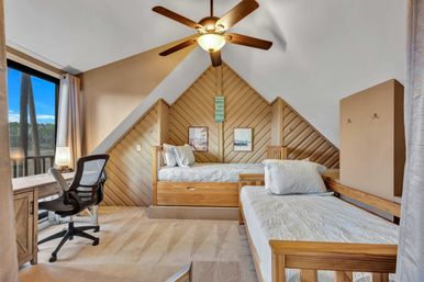 Cozy attic-style bedroom with sloped wood-paneled accent wall, two twin wooden beds with white quilts, ceiling fan, study desk and ergonomic chair by a large window overlooking pine trees, neutral carpet and soft natural light.