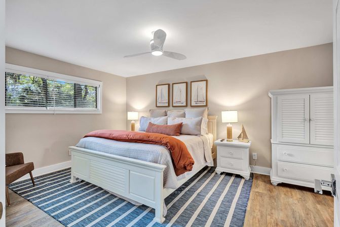 Bright coastal-style bedroom with white wood bed and armoire, navy-striped rug, rust-orange throw, bedside lamps and ceiling fan over hardwood floors.