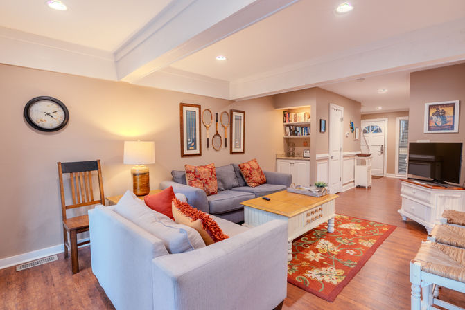 Cozy open living room in a suburban home — light-blue sofas with red patterned pillows, wooden coffee table on a floral red area rug, built-in bookshelves and entry door in the background.