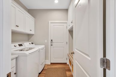Bright modern home laundry room with white top-load washer and dryer, overhead white cabinets, hardwood floors, woven bench storage with wicker baskets, and a white door with electronic lock.