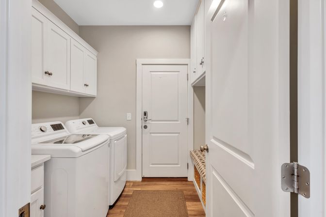Bright modern home laundry room with white top-load washer and dryer, overhead white cabinets, hardwood floors, woven bench storage with wicker baskets, and a white door with electronic lock.