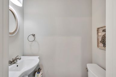 Cozy bright powder room with white pedestal sink, chrome faucet and towel ring, round rustic wood-framed mirror, white toilet, and nautical rope wall art on neutral gray walls.