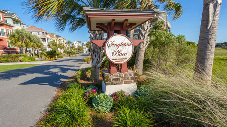 Entrance sign for a private oceanfront community framed by palm trees, coastal-style colorful homes along a sunny street, and landscaped grasses and flowering plants.