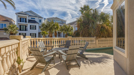 Sunlit coastal poolside patio with gray Adirondack chairs around a round table, decorative stone balustrade, palm trees and multi-story beach houses under a blue sky