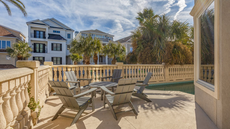 Sunlit coastal poolside patio with gray Adirondack chairs around a round table, decorative stone balustrade, palm trees and multi-story beach houses under a blue sky