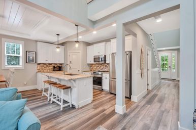 Sunny coastal-style open-concept kitchen in a beach cottage with white cabinets, marble-topped island and three wooden stools, pendant lights, stainless steel appliances, mosaic tile backsplash, and wood-look plank floors leading to a bright entryway.