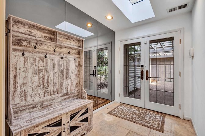 Bright home entryway mudroom with distressed whitewashed wooden bench and hooks, floor-to-ceiling mirror, skylight, patterned rug, tile floor, and glass French doors opening to a patio.