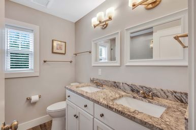 Bright modern residential bathroom with double vanity and veined stone countertop, white cabinets, undermount sinks, brass fixtures and light sconces, framed mirrors, toilet and window shutters