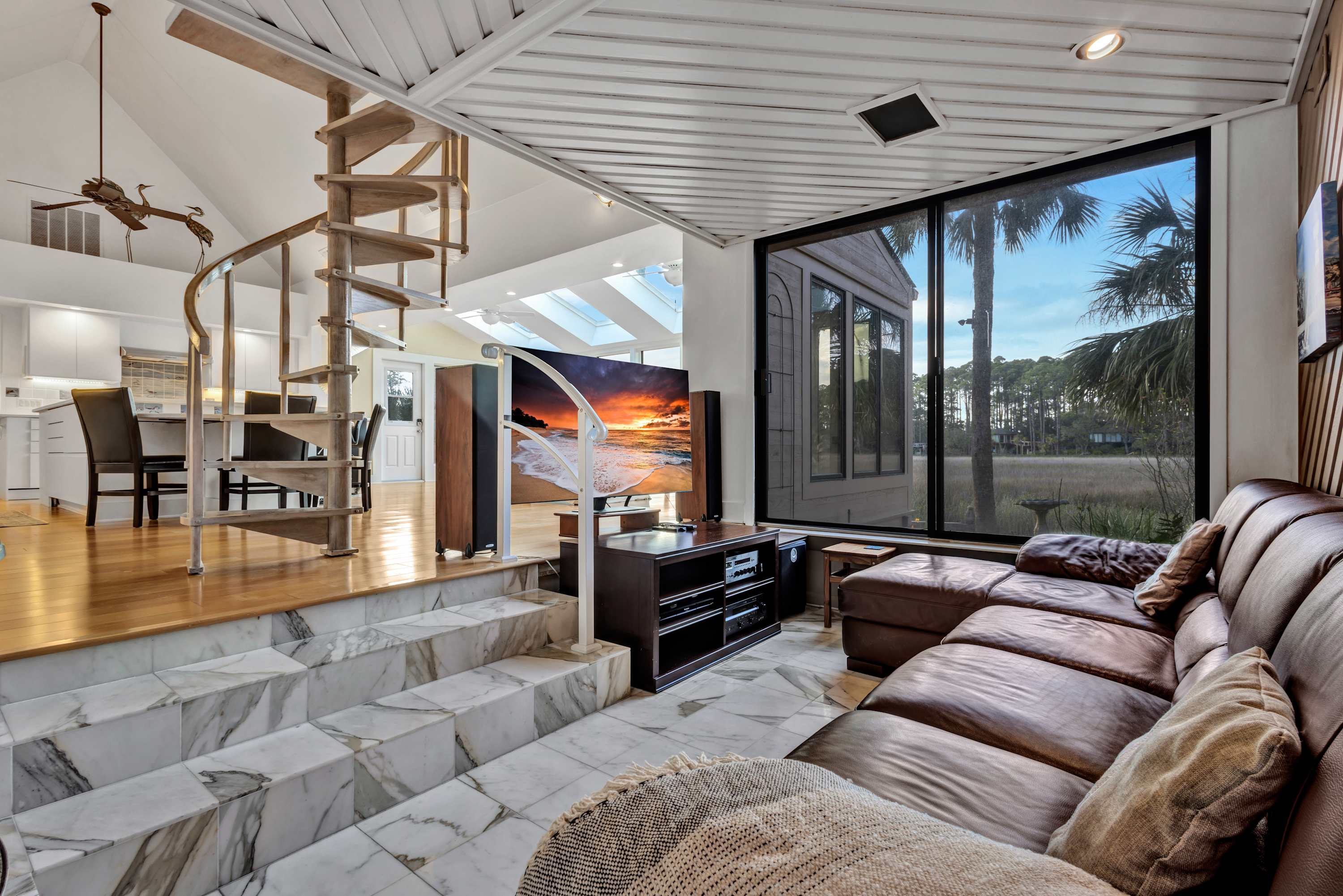 Sunlit modern coastal living room with brown leather sectional, marble sunken seating area and steps, spiral staircase to an open-plan kitchen, large window framing palm trees and marsh, and a TV displaying a beach sunset.