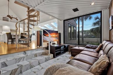 Sunlit modern coastal living room with brown leather sectional, marble sunken seating area and steps, spiral staircase to an open-plan kitchen, large window framing palm trees and marsh, and a TV displaying a beach sunset.