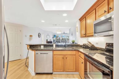 Bright open-plan kitchen with black granite countertops, maple cabinets, stainless steel appliances, center sink island under a skylight, hardwood floors and balcony with palm-tree view.